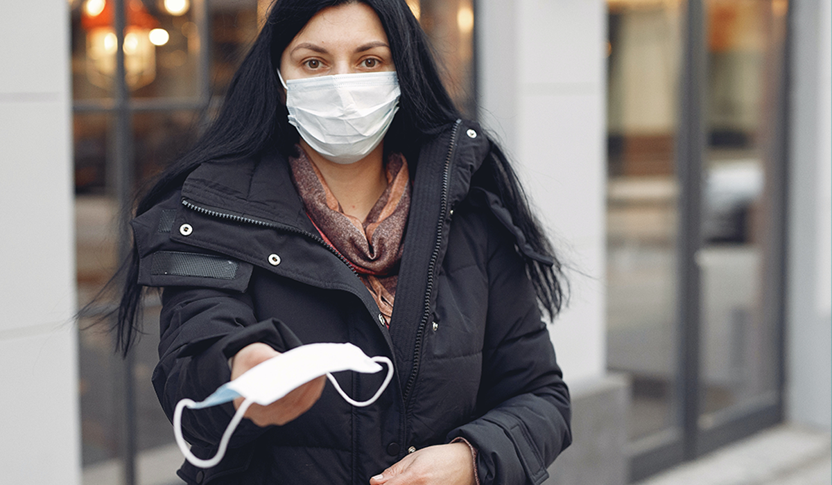 Woman with dark hair and black coat wearing a face mask offers a face mask to a passerby