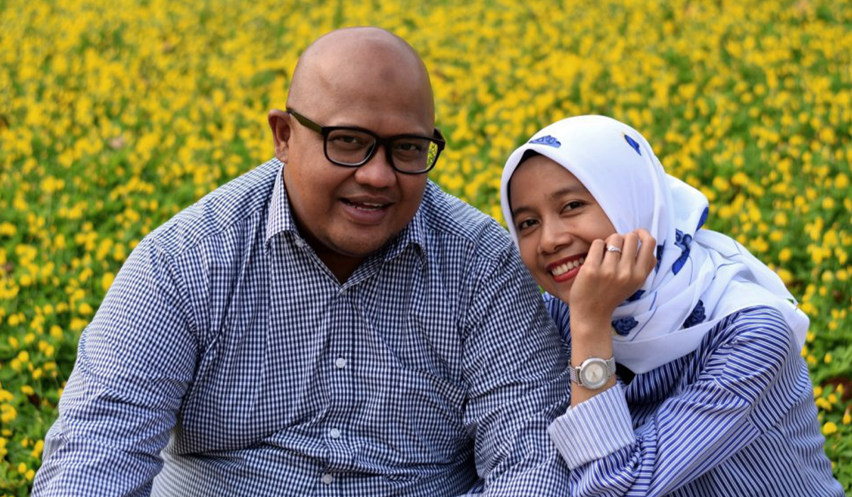 Man in glasses and woman in hijab sit in a field and smile at the camera