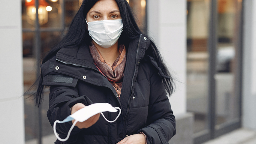 Woman with dark hair and black coat offers face mask to passerby